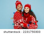 Side view merry young couple two man woman wear red Christmas sweater Santa hat wrapped in garlands posing hug cuddle isolated on plain pastel light blue background Happy New Year 2023 holiday concept