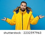 Young caucasian man 20s wear yellow down jacket spread hands shrugging shoulders looking puzzled, have no idea isolated on plain blue color background studio portrait. People winter lifestyle concept