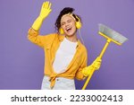Small photo of Young housekeeper woman wear yellow shirt rubber gloves headphones listen music dance tidy up hold broom sweeps floor clean house isolated on plain pastel purple background studio. Housework concept