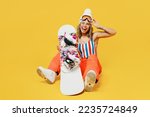 Snowboarder cheerful excited fun woman wear orange ski suit mask hat swimsuit spend extreme weekend sitting hold goggles wink isolated on plain yellow background. Winter sport hobby trip relax concept