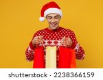 Young fun satisfied man wear red knitted christmas sweater Santa hat hold open look at shopping paper package bags isolated on plain yellow background. Black Friday New Year 2023 sale buy day concept