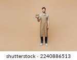 Small photo of Full body young happy man barista barman employee wear brown apron work in shop hold craft paper brown cup coffee to go isolated on plain pastel light beige background. Small business startup concept