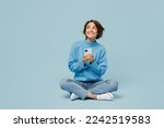 Full body fun young woman wear knitted sweater hold in hand use mobile cell phone look aside on workspace isolated on plain pastel light blue cyan background studio portrait. People lifestyle concept
