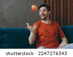 Small photo of Young happy satisfied man wears red t-shirt eat toss up apple sit on blue sofa couch stay at home hotel flat rest relax spend free spare time in living room indoors grey wall. People lounge concept