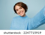 Close up young caucasian woman wear knitted sweater look camera doing selfie shot pov on mobile cell phone isolated on plain pastel light blue cyan background studio portrait. People lifestyle concept