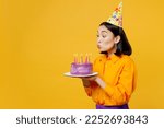 SIde view happy fun young woman wear casual clothes hat celebrate hold purple cake blow out candles make wish in special moment isolated on plain yellow background. Birthday 8 14 holiday party concept