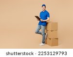 Full body delivery guy employee man wears blue cap t-shirt uniform workwear work as dealer courier stand near stack cardboard boxes hold clipboard papers documents isolated on plain beige background