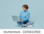Full body young smiling happy fun cool IT woman wear knitted sweater hold use work on laptop pc computer isolated on plain pastel light blue cyan background studio portrait. People lifestyle concept