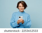 Young smiling happy fun caucasian woman wear knitted sweater hold in hand use mobile cell phone chatting isolated on plain pastel light blue cyan background studio portrait. People lifestyle concept