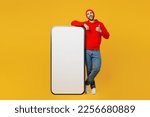 Full body happy young man wear red hoody hat near big huge blank screen mobile cell phone smartphone with workspace copy space mockup area showing thumb up isolated on plain yellow color background