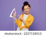 Small photo of Young satisfied happy fun housekeeper woman wear yellow shirt tidy up hold in hands point index finger on iron isolated on plain pastel light purple background studio. Housework housekeeping concept