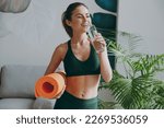 Young strong sporty athletic fitness trainer instructor woman wearing green tracksuit holding in hand yoga mat drink water training do exercises at home gym indoor. Workout sport motivation concept