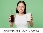 Young woman wearing white clothes hold in hands bottle of soda pop fizzy cola give glass of milk isolated on plain pastel green background. Proper nutrition healthy fast food unhealthy choice concept