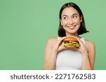 Young happy woman wear white clothes hold eat burger look aside on workspace area mockup isolated on plain pastel light green background. Proper nutrition healthy fast food unhealthy choice concept