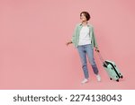 Full body side view young traveler woman wear green shirt hold suitcase isolated on plain pastel light pink background Tourist travel abroad in free time rest getaway Air flight trip journey concept