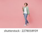 Full body side view young woman 20s she wear green shirt white t-shirt hold takeaway delivery craft paper brown cup coffee to go isolated on plain pastel light pink background People lifestyle concept