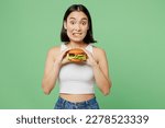 Small photo of Young surprised pop-eyed look camera woman wear white clothes holding eat burger isolated on plain pastel light green background. Proper nutrition healthy lifestyle fast food unhealthy choice concept
