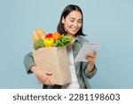 Young happy woman wear casual clothes hold brown paper bag with food products read liat of ingredients isolated on plain blue cyan background studio portrait. Delivery service from shop or restaurant