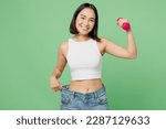 Small photo of Young woman wears white clothes show loose pants on waist after weightloss hold dumbbell isolated on plain pastel light green background. Proper nutrition healthy fast food unhealthy choice concept
