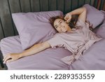 Small photo of Young happy woman wear purple t-shirt pajama lying in bed look aside rest relax spend time in bedroom lounge home in own room hotel wake up dream be lost in reverie good mood day. Real estate concept