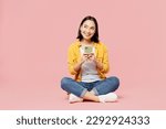 Full body young woman of Asian ethnicity wear yellow shirt white t-shirt sitting hold in hand use mobile cell phone isolated on plain pastel light pink background studio portrait. Lifestyle concept