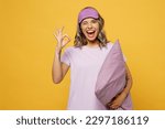 Small photo of Calm young woman she wears purple pyjamas jam sleep eye mask rest relax at home show ok okay gesture hold pillow wink isolated on plain yellow background studio portrait. Good mood night nap concept