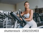 Dreamful minded happy young strong sporty athletic sportswoman woman 20s in white sportswear earphones listen music look aside warm up training use exercise bike in gym indoor Workout sport concept.