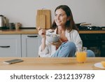 Young fun smiling european happy housewife woman wear casual clothes look aside eat breakfast muesli cereals with milk fruit in bowl cooking food in light kitchen at home alone. Healthy diet concept