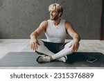 Full body young sporty athletic sportsman man wear white tank shirt black shorts use mobile cell phone listen music in earphones look aside sit on caremat warm up train indoor at gym Workout concept