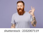Small photo of Young cheerful fun redhead bearded man wearing violet t-shirt casual clothes showing okay ok gesture wink blink eye isolated on plain pastel light purple background studio portrait. Lifestyle concept