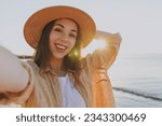 Close up smiling young woman 20s in straw hat shirt summer casual clothes do selfie shot mobile cell phone outdoors at sunrise sun dawn over sea background People vacation lifestyle journey concept.