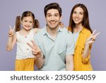 Young happy parents mom dad with child kid daughter girl 6 years old wears blue yellow casual clothes look camera show victory gesture v-sign isolated on plain purple background. Family day concept