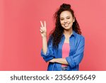 Young smiling happy cheerful woman of African American ethnicity she wear blue shirt casual clothes showing victory sign look camera isolated on plain pastel pink background studio. Lifestyle concept