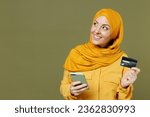 Small photo of Wistful young arabian asian muslim woman in abaya hijab yellow clothes hold mobile cell phone credit bank card look aside isolated on olive green background studio. People uae islam religious concept