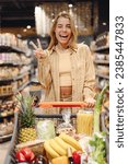 Small photo of Young cool customer woman wear casual clothes show v-sign wink shopping at supermaket store grocery shop buying with trolley cart choose products inside hypermarket. Purchasing food gastronomy concept