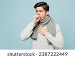 Small photo of Young ill sick man wear gray sweater scarf cough sneeze cover mouth with hand isolated on plain blue background studio portrait. Healthy lifestyle disease virus treatment cold season recovery concept