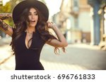 Portrait of gorgeous glam tattooed lady with long wavy hair and beautiful make up holding her trendy fedora hat for it not to be blown away by the wind. Her mouth open with surprise. Copy-space
