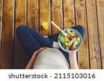 View from above of pregnant woman with fresh salad bowl and orange juice, expectant mother having healthy lunch while sitting on wooden floor in lotus pose, cropped. Nutrition during pregnancy