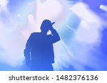 Small photo of Stock photo of young rap singer with mic in hand singing popular song on stage in blue lights.Hip hop artist performing live on scene in music hall.Repper with microphone in royalty free backgrounds