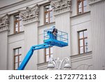 Small photo of Construction worker in lift bucket of crane restore and repair historic facade of building. Man in cradle, restoring plaster decoration on facade. Workers painting building, renovating and repair work