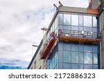 Small photo of Suspended platform, construction cradle hanging on building. Installation and repair work. Building facades construction works. Glass facade of high-rise building under construction, finishing works
