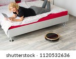 A middle aged woman is reading on her digital tablet on her bed while her vaccum cleaning robot is cleaning the wooden floor at home. Household, leisure and technology concept
