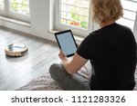 A middle aged woman is reading on her digital tablet on her bed while her vaccum cleaning robot is cleaning the wooden floor at home. Blank screen with space for your text, website or message on the