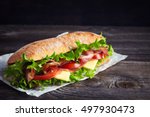 Fresh submarine sandwich with ham, cheese, bacon, tomatoes, cucumbers, lettuce and onions on dark wooden background