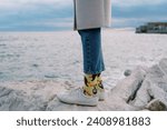 Women legs in sneakers and jeans stand on a stone by the sea. Cropped. Faceless