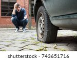Small photo of Crouched Worried Young Man With Hand On Head Pointing At Punctured Car Tire