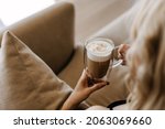 Closeup of a woman holding a double glass cup of coffee with foam, sitting on sofa at home.