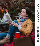 Small photo of young woman brown coat sits chair at table reading book with plaid thrown over her head open air against autumn reddened foliage. autumn fashion season. Rest, tranquility, backyard, patio, melancholy