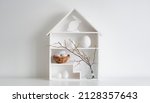 Small photo of springtime Home minimalist interior with Easter decor. Branches with budding buds in a glass vase on shelves in the shape of a house, Easter eggs in a nest basket on a white background.