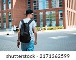 young man fashionable stylish guy sunglasses black cap walking around city holding a smartphone his hand and listening music headphones. Tourism, summer vacation, rear view, back view, backpack